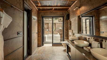 Bathroom of the Legacy Suite With Jacuzzi with stone-textured walls, double vessel sinks, wooden ceiling beams, and glass doors leading to a jacuzzi tub area at AamaGhati Wildlife Resort.