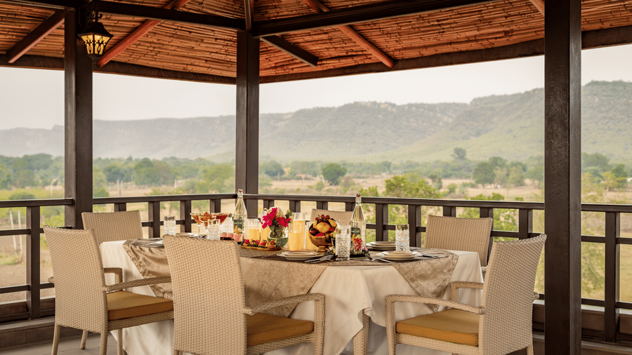 Covered outdoor dining terrace of Badal Mahal under a thatched roof with a set dining table overlooking distant green hills at AamaGhati Wildlife Resort.
