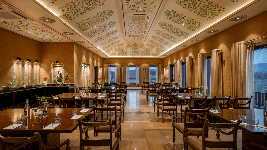 Elegant indoor dining hall of Baradari at AamaGhati Wildlife Resort with painted floral ceilings, earthen wall finishes, and neatly set wooden dining tables.