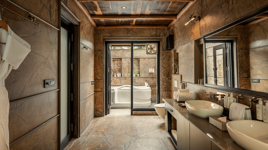 Bathroom of the Legacy Suite With Jacuzzi with stone-textured walls, double vessel sinks, wooden ceiling beams, and glass doors leading to a jacuzzi tub area at AamaGhati Wildlife Resort.