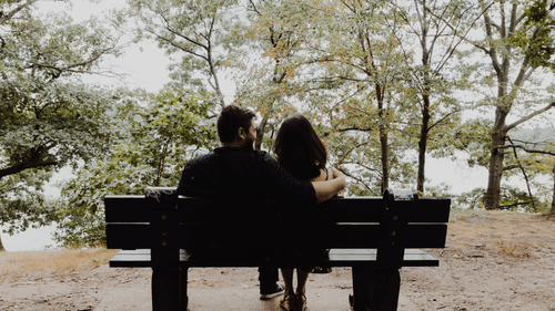 A silhouette of two people sitting on a bench with trees in the background.