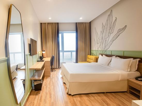 A side wide-angle view of the Superior Deluxe room at Hotel Bawa Regency, Dadar, featuring a comfy bed, a large hexagon-shaped mirror, a flat-screen TV, and a balcony.