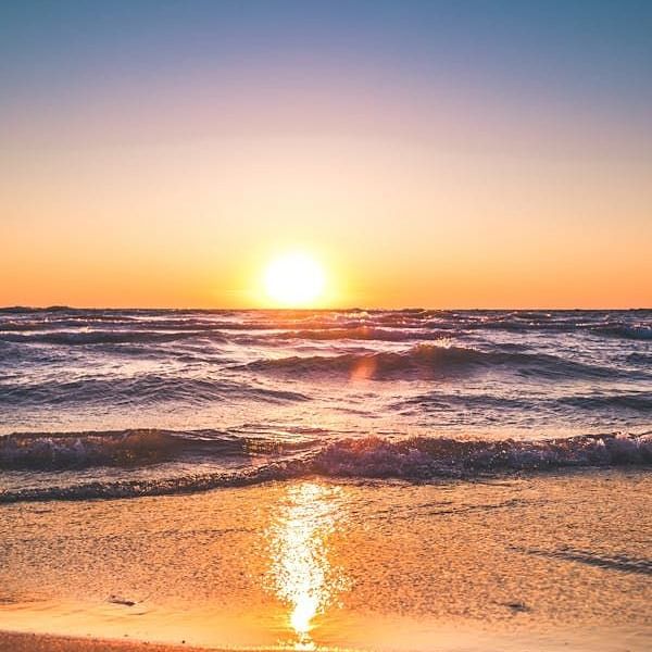 The sun sets over the ocean, casting a bright glow on the horizon, waves, and sandy shore.
