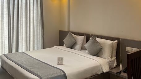 Focus view of the Daksh Bliss room at Daksh The Valley Resort, Saputara, showcasing its queen size bed, large windows with closed curtains, and warm overhead lighting.