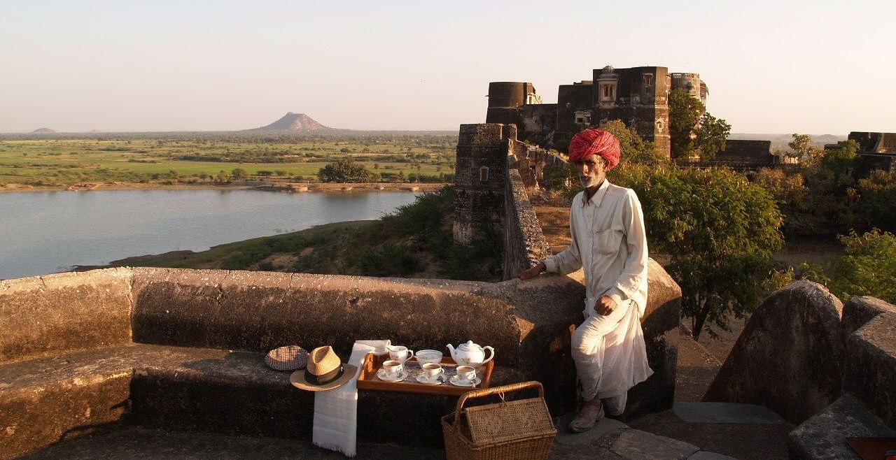 A man in traditional attire sits on a stone wall overlooking a landscape with a body of water, with a tea set laid out on a table beside him- Shahpura Bagh.