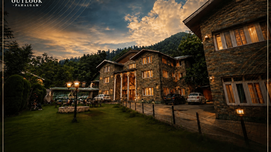 Exterior view of Hotel Outlook, Pahalgam, showing stone buildings next to a lawn, illuminated under a cloudy sky with hills in the background.