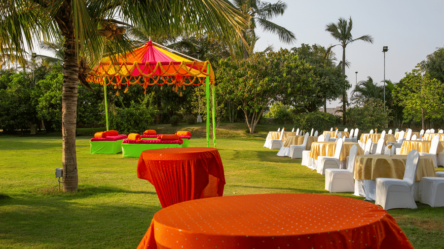 An outdoor dining area with orange-draped tables on a green lawn with palm trees and a vibrant tent - Grande Bay Resort & Spa, Mamallapuram