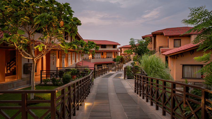 A raised wooden deck pathway bordered by dark timber railings, soft ground lighting, and terracotta-roofed cottages at AamaGhati Wildlife Resort.