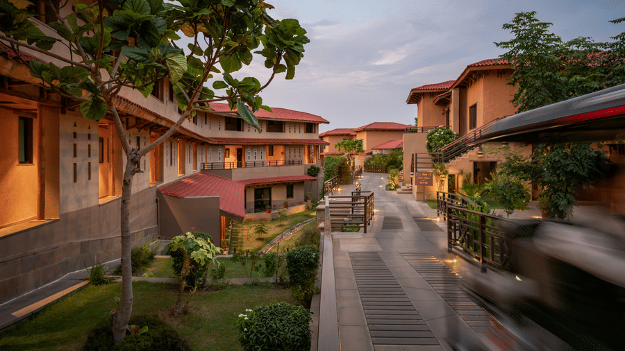A raised wooden deck pathway bordered by dark timber railings, soft ground lighting, and guests walking by in the foreground at AamaGhati Wildlife Resort.