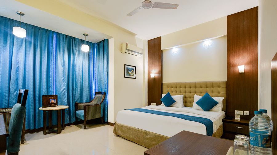 The Pamposh, New Delhi | A room with tables, a chair, a large bed, sporeads, pillows, windows covered with curtains, and doors.
