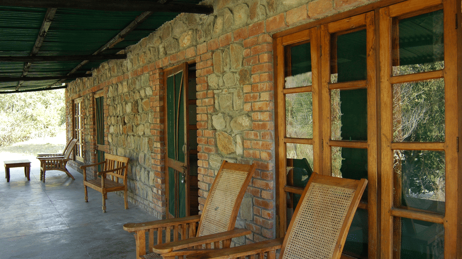 A shaded covered veranda of a rustic stone cottage furnished with wooden lounge chairs and glass doors.