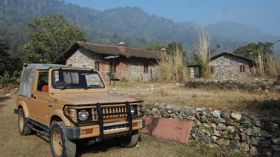 A beige safari jeep parked on a dirt path in front of stone cottages with misty mountains in the background.