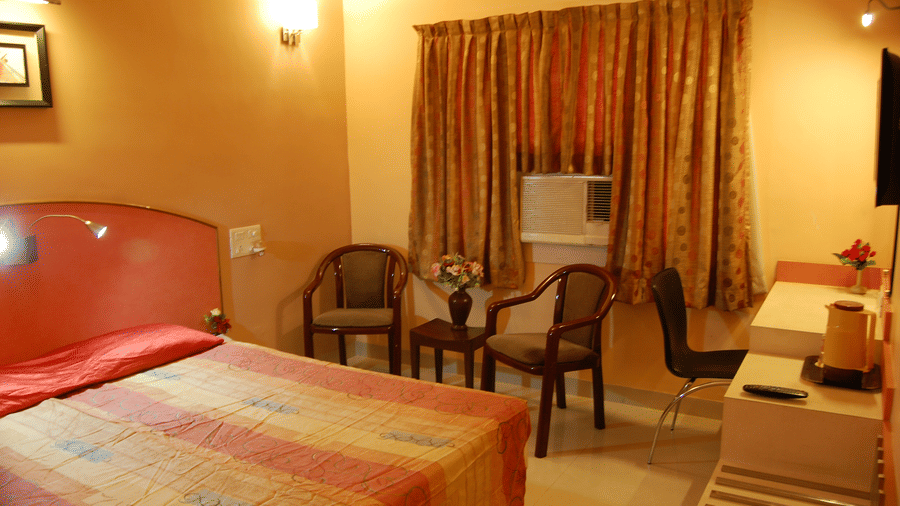A cosy hotel bedroom with a double bed, warm peach-coloured walls, wooden chairs, and an air-conditioning unit - Hotel Pandian