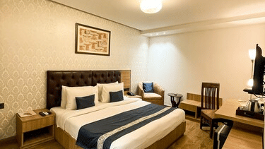 A clean and inviting double room featuring a dark tufted headboard, blue bed runner, and warm ambient ceiling lighting - Hotel 91 HUDA City Centre, Gurugram