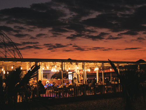 An illuminated outdoor structure filled with people, under a dark, cloudy sky with a sunset glow visible on the horizon.