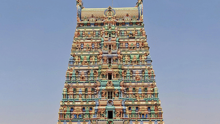 A tall gopuram temple tower covered in multicoloured sculpted figures, photographed from ground level with surrounding buildings at the bottom.
