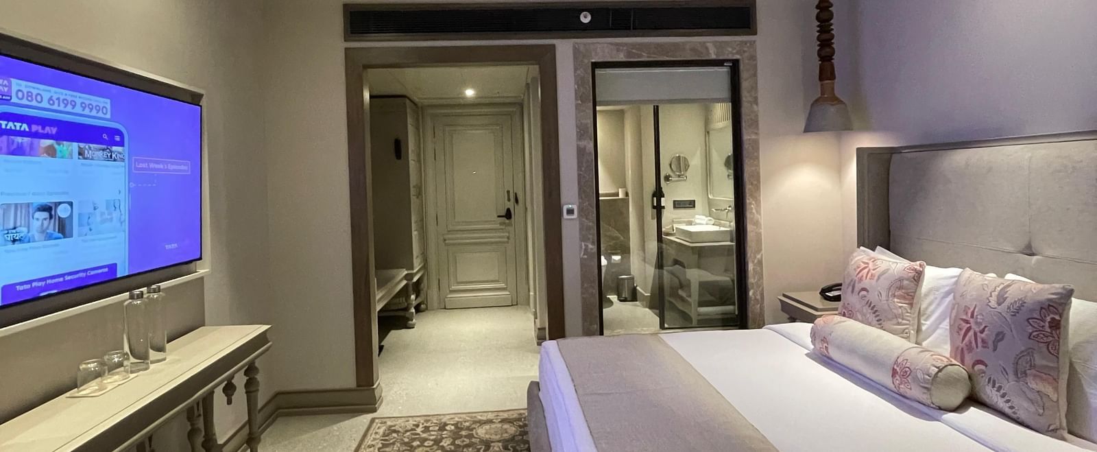 A wide-angle view of the bedroom at The Evren, Vagator, showing the bed, television, and the open entryway leading to the bathroom. A patterned rug covers a portion of the floor.