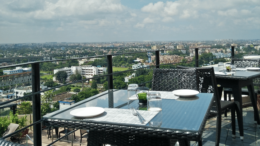 A dining table with a glass top situated besides a railing overlooking the beautiful landscape of Kolkata - Senses Hotel