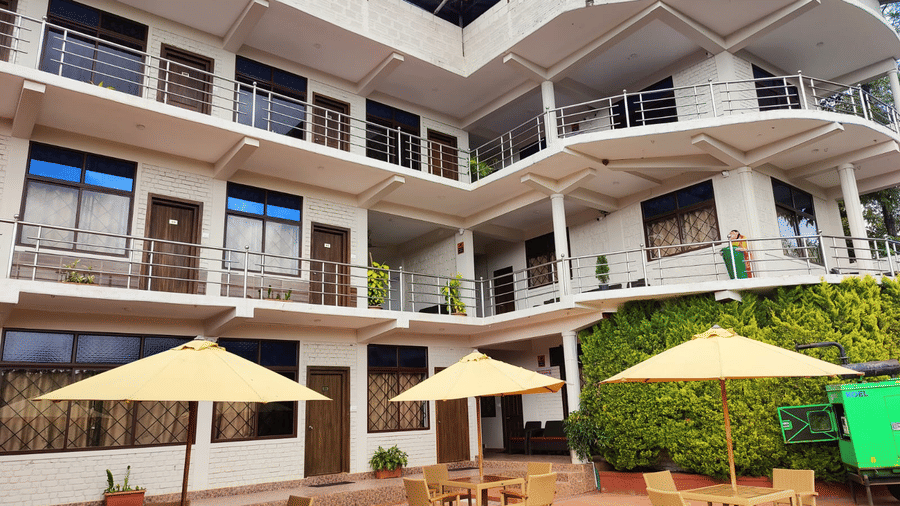 Facade of Woodstock Resorts, Coorg, featuring the resort building, patio umbrellas, chairs and tables, and a hedge for decoration.