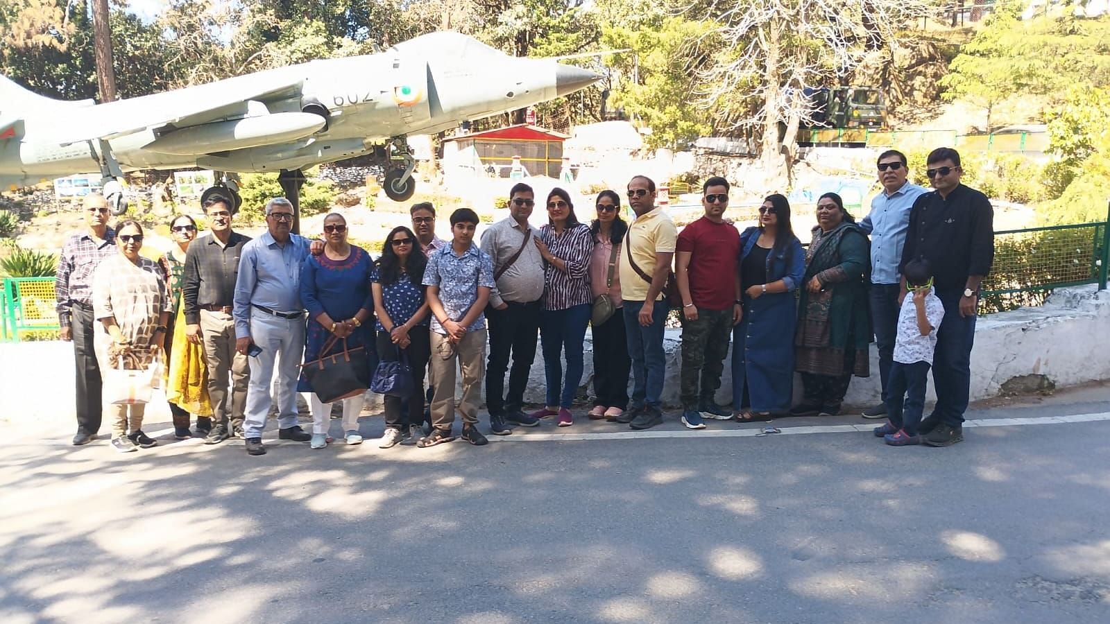 A large group of people standing together for a group photo in front of a fighter aircraft display in an outdoor setting surrounded by trees.