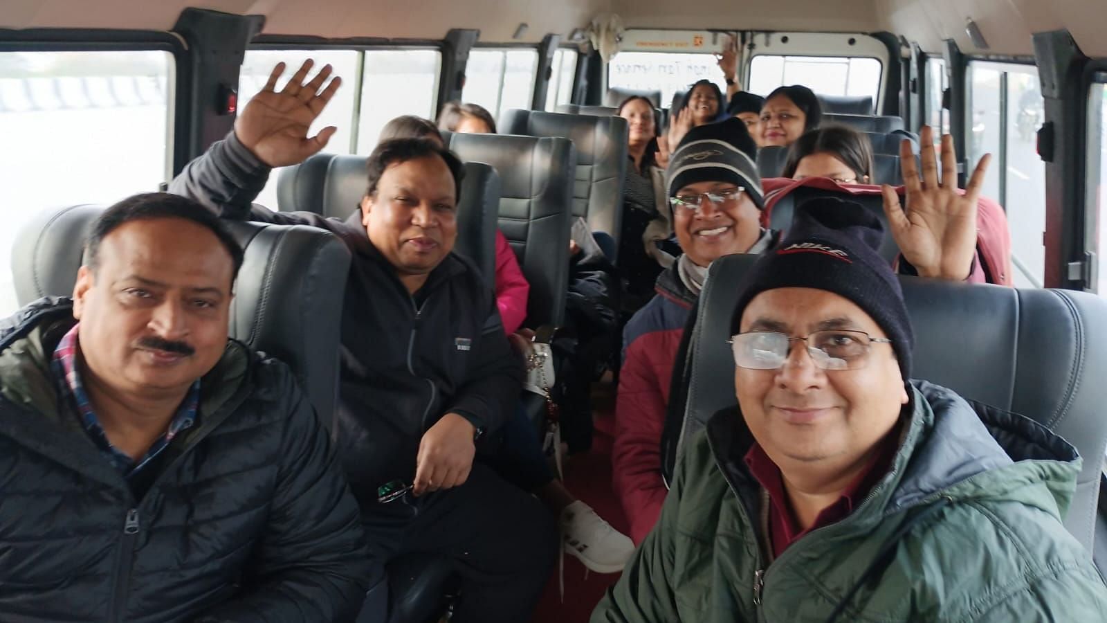 A group of people sitting inside a van, smiling and waving at the camera. The image shows individuals dressed in warm clothing, with some wearing glasses and hats.
