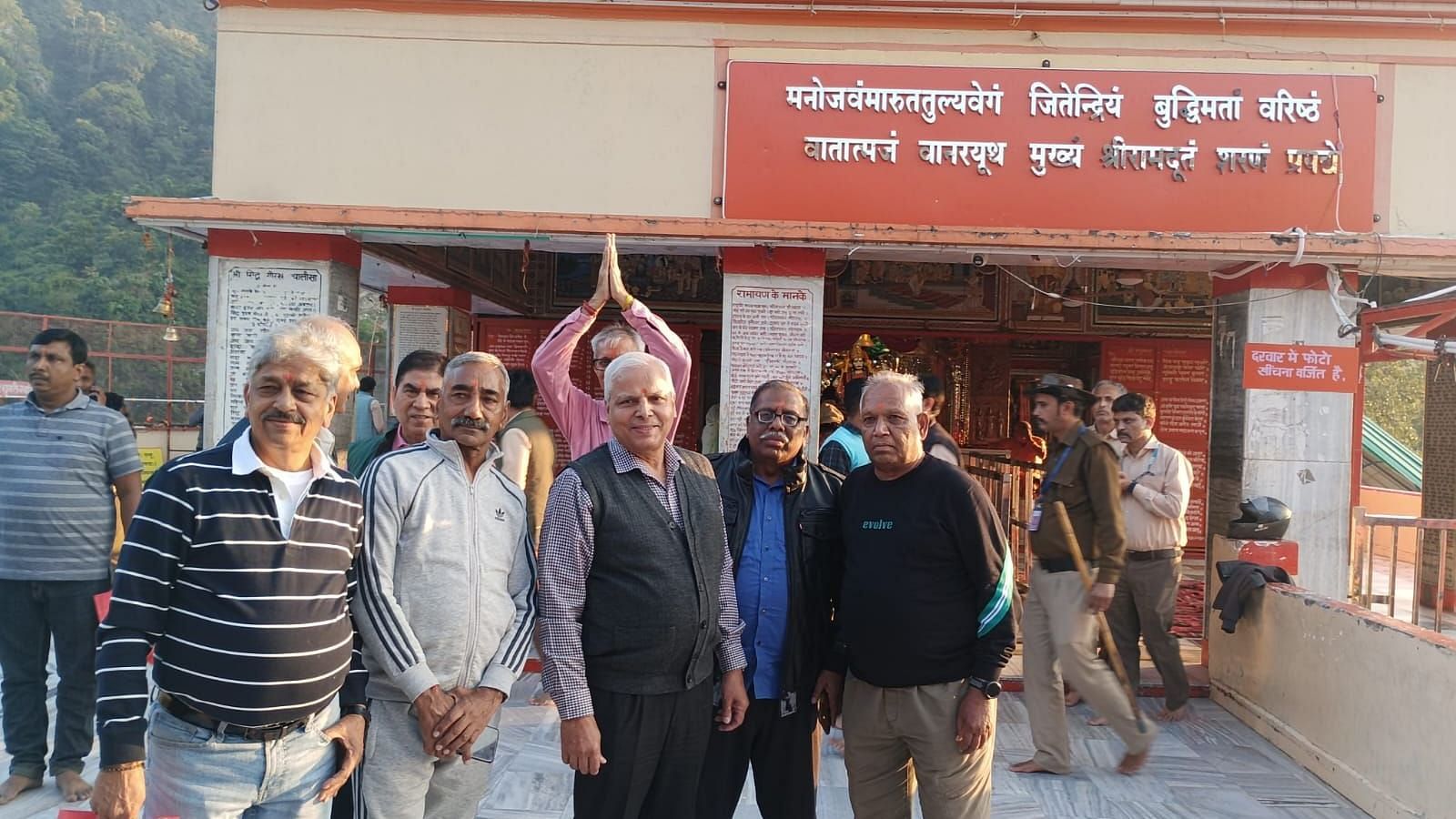 Group of elderly men posing for a photo in front of the entrance to the Shri Siddhbali Dham temple in Kotdwar, Uttarakhand, with a mountainous forest backdrop. The temple signboard with Hindi script and the idol of Lord Hanuman is visible in the background