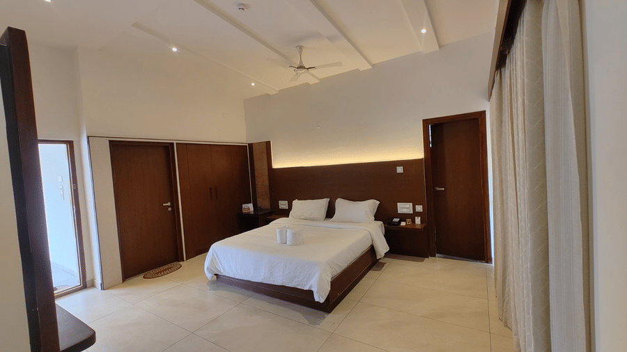 A well-lit hotel bedroom featuring a double bed with white linens, wooden wall panels, and a ceiling fan | Lords Sahyadri Resort, Shivamogga