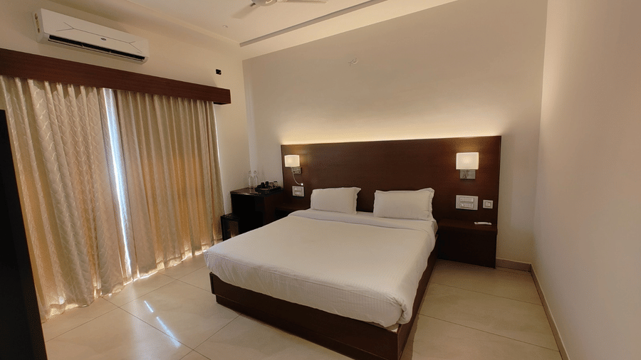 A hotel bedroom featuring a double bed, a dark wooden headboard with built-in side lights, and large glass doors with floor-to-length curtains | Lords Sahyadri Resort, Shivamogga