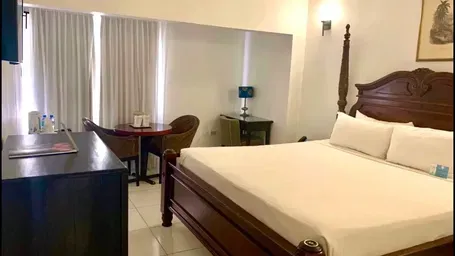 A well-lit hotel room features a large wooden bed with white linens, a dark dresser, and a seating area near a window | Executive King Room | Altamont Court, Jamaica