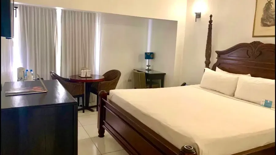 A well-lit hotel room features a large wooden bed with white linens, a dark dresser, and a seating area near a window | Executive King Room | Altamont Court, Jamaica