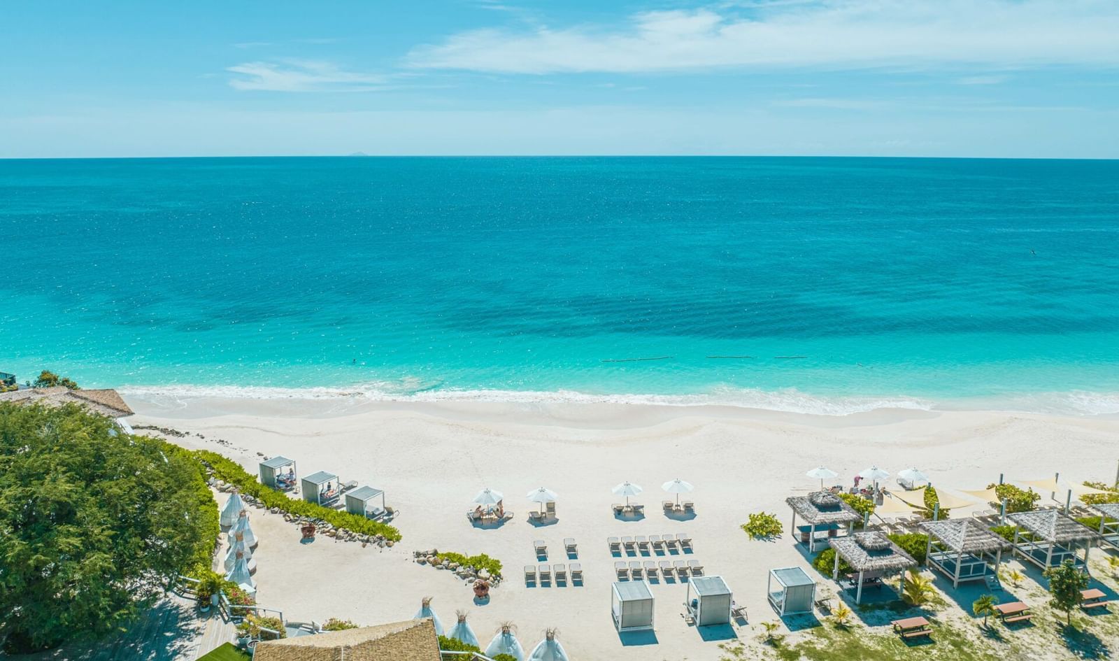 An aerial view of a resort beach with white sand, blue ocean, numerous lounge setups, and thatched structures.