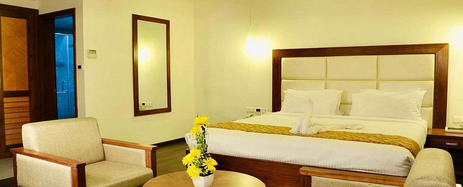 A close up view of well-lit Suite at Dhanalakshmi Srinivasan Hotels, Perambalur featuring a king size bed, two armchairs, a small table, and a work desk with a chair.