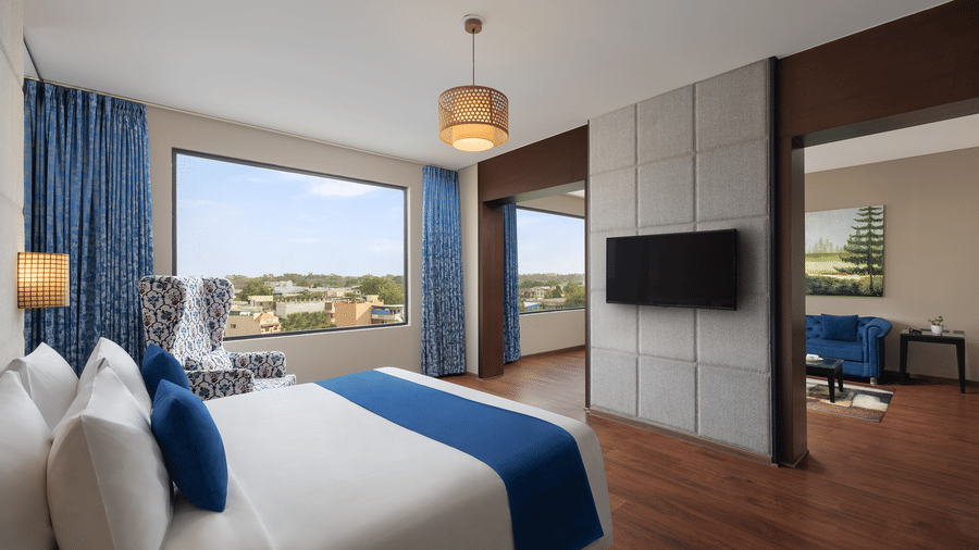 Deluxe Suite at Essentia Luxury Hotel, Indore, with a double bed in front of a TV on the wall next to a large window and a separate living area behind the wall