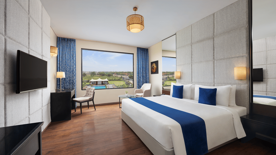 One of the best Premium Rooms in Indore at Essentia Luxury Hotel featuring a double bed, a television, work desk and chair, and an expansive window overlooking the green landscape.