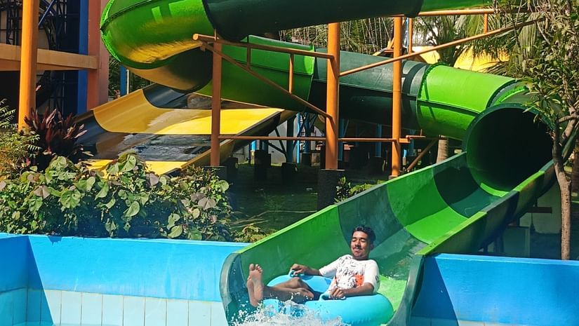 A rider splashes into the pool at the end of a green water slide, surrounded by palm trees and mountains.