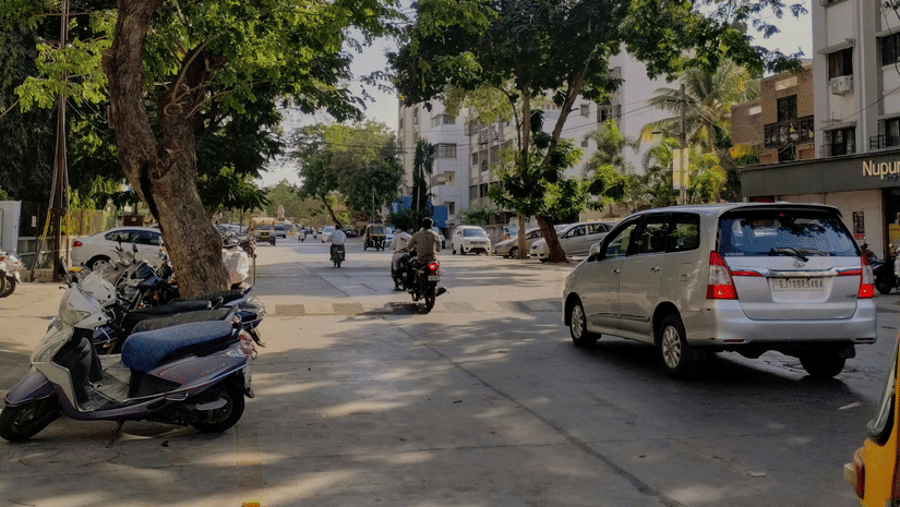 A Jamnagar street with trees, parked vehicles, and people walking along the roadside.
