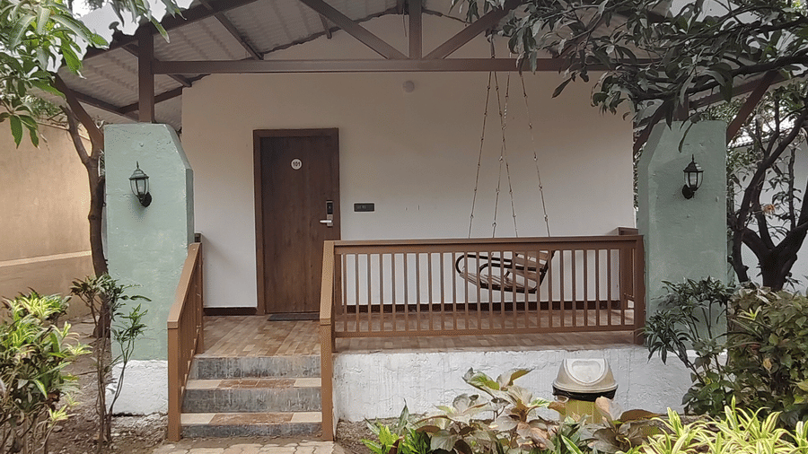 Resort cottage with cobblestone pathway, manicured garden, globe light, hanging porch swing, and ornate gabled roof amid green trees.