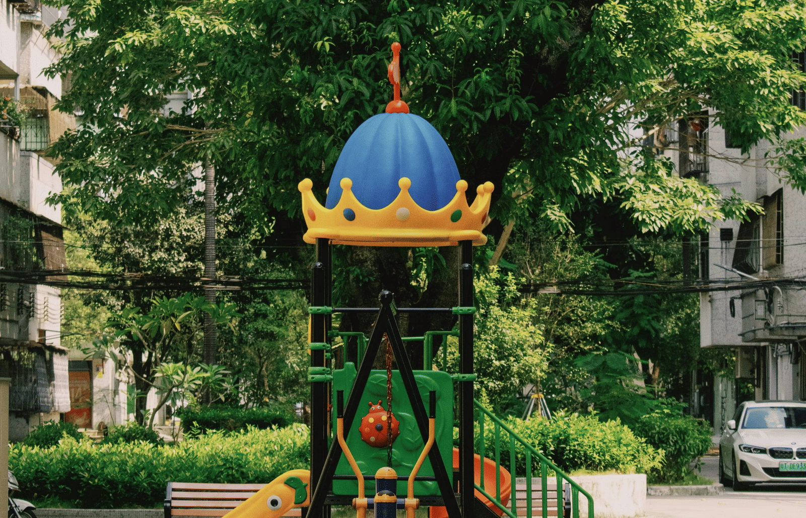 A colourful playground with a blue dome is centred in the frame, surrounded by lush green trees and some apartment buildings in the background. 