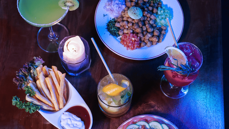 Overhead shot of vibrant food dishes and drinks on a dark wooden table, illuminated by blue and red lighting at Hotel Hukam's Lalit Mahal.