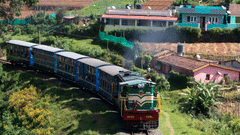 Nilgiri Mountain Railway featuring the toy train travelling along a hillside railway, passing lush vegetation, terraced slopes, and scattered buildings in the distance