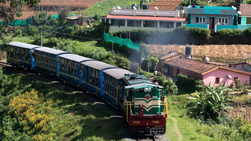 Nilgiri Mountain Railway featuring the toy train travelling along a hillside railway, passing lush vegetation, terraced slopes, and scattered buildings in the distance
