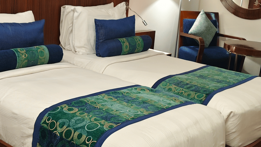 A vertical view of the two single beds inside the Superior Twin Room at Pride Premier Chennai. Artwork hangs on the wall behind the headboards, and a lamp stands near a window seating area in the background.