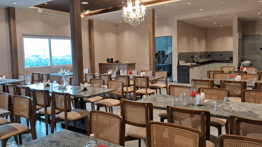 Multiple wooden dining tables and cane-back chairs, arranged in a restaurant lit by ceiling chandeliers at Savyasaachii Hotel.