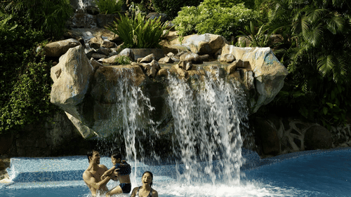 A waterfall flowing over rocks into a swimming pool with surrounding tropical vegetation and people in the pool at he Retreat Hotel and Convention Centre, one of the best hotels in Mumbai.