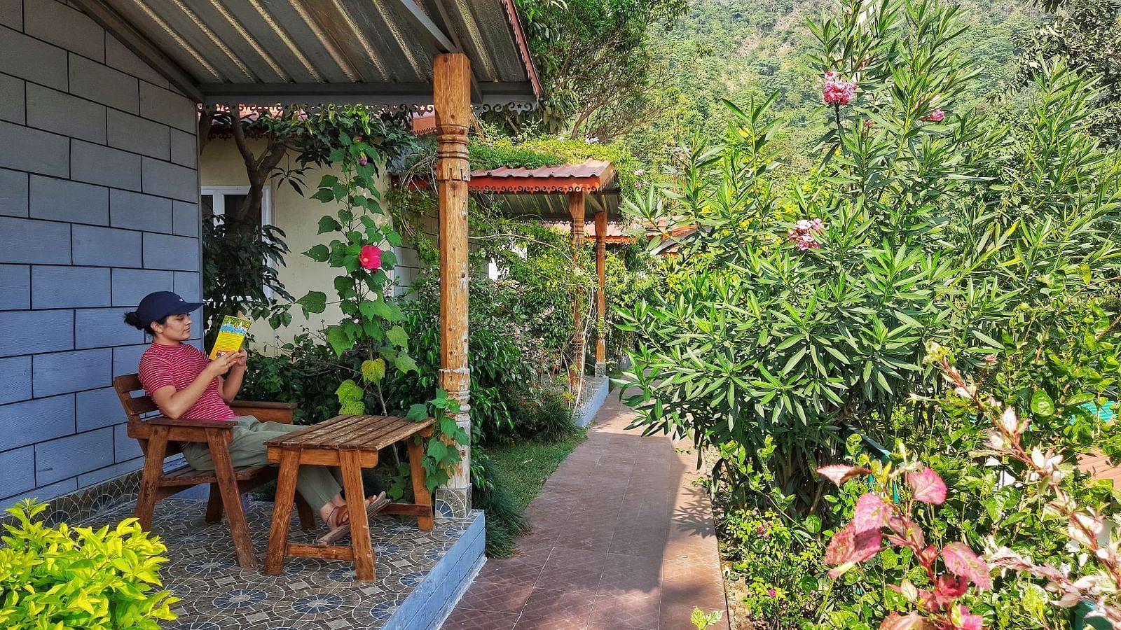 a person reading a book outside one of the room with a garden in front of them - Neer Ganga Resorts