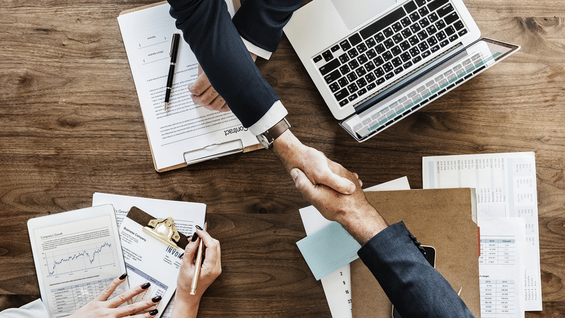 two people shaking their hands above a table with laptop and papers on it