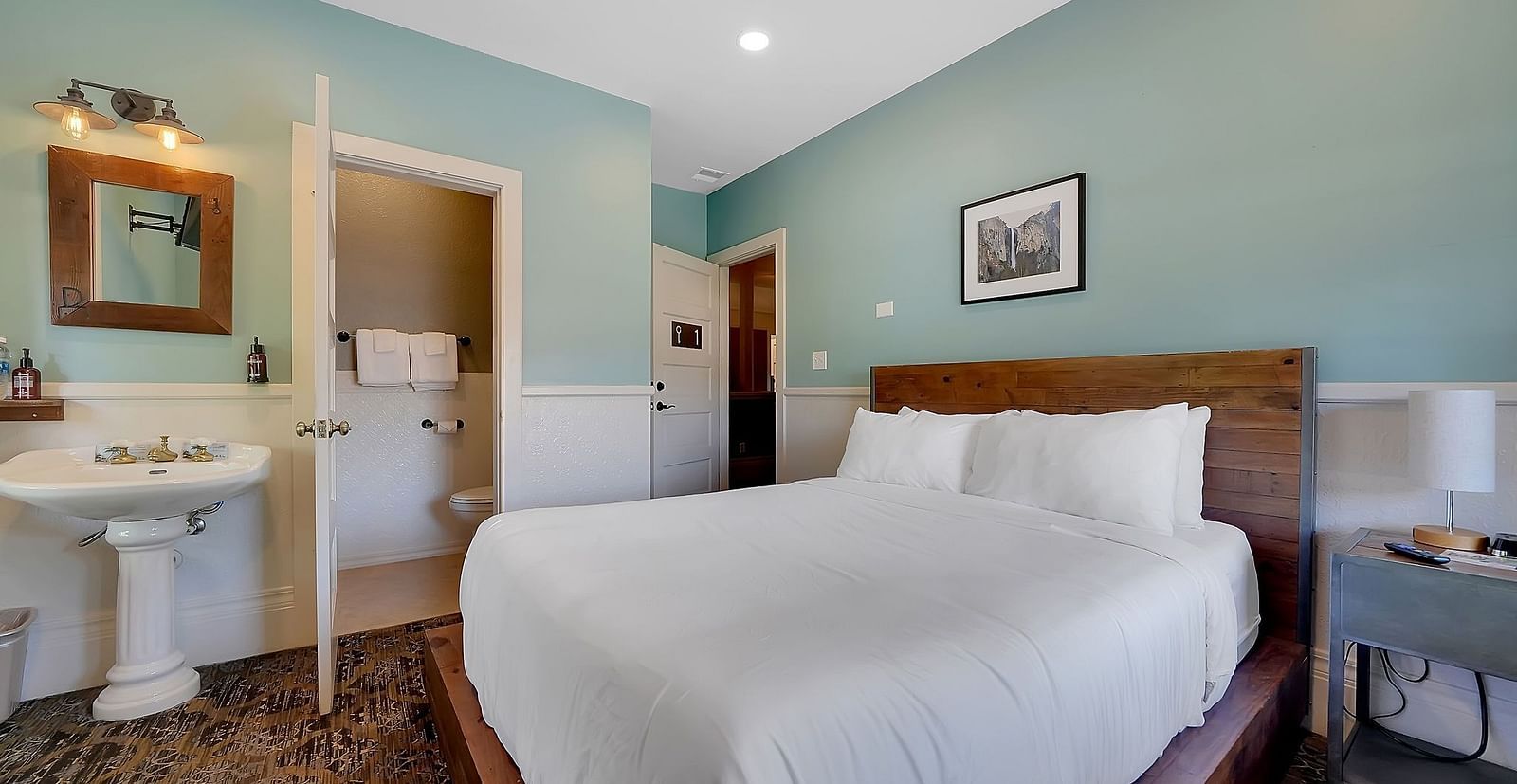 The Groveland Hotel's compact room features sage green walls, a wooden bed with crisp white bedding, and a pedestal sink with mirror.