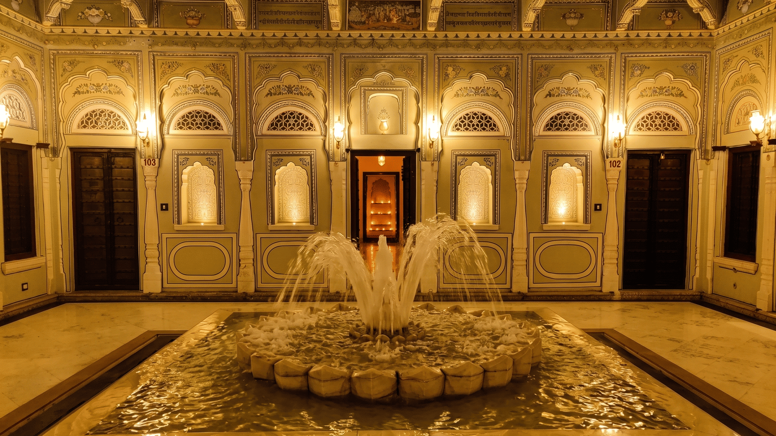 Courtyard at night with a marble lotus fountain, lamp-lit arched niches and painted walls with guest room doors at BluSalzz Vintage - Harlalka Haveli.