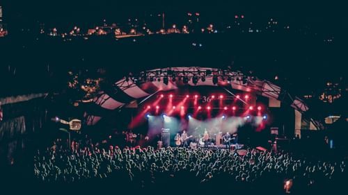 A wide-angle view of a nighttime outdoor concert with a large crowd and a stage lit by vibrant pink and purple lights.