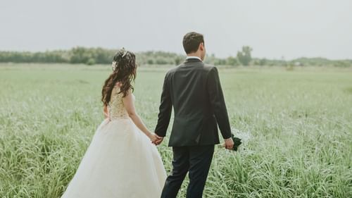 A bride and groom in wedding attires walking thorugh a field while holding hands